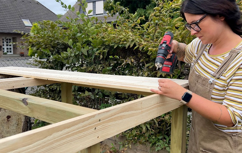 A woman uses a cordless drill to attach wooden boards to the platform structure.