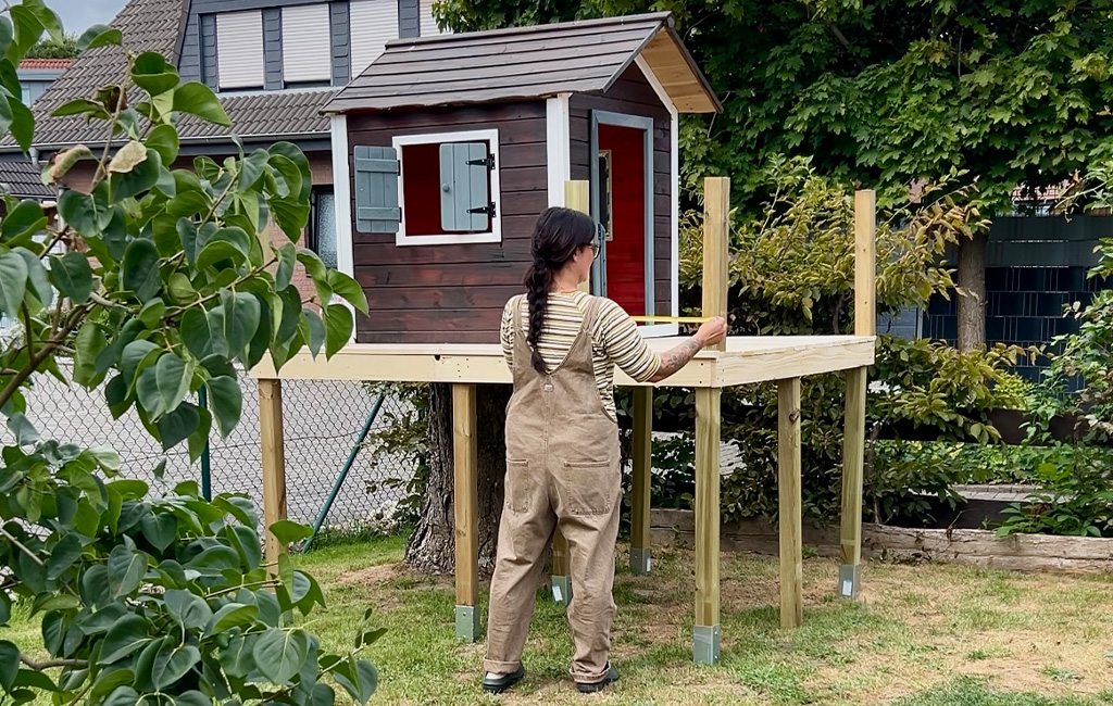 A woman measures the railing of the elevated platform in front of a partially assembled playhouse.