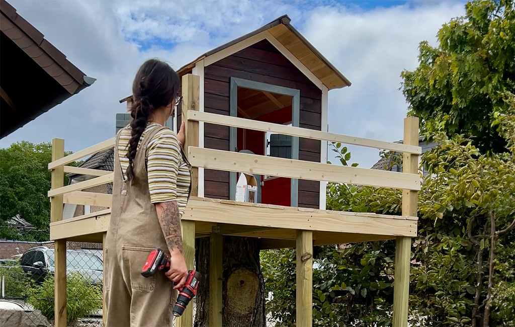 A woman stands with a cordless drill in front of the half-finished playhouse with a mounted railing.