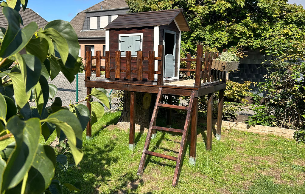 The finished playhouse with dark-stained wooden railing stands elevated on stilts in the garden.