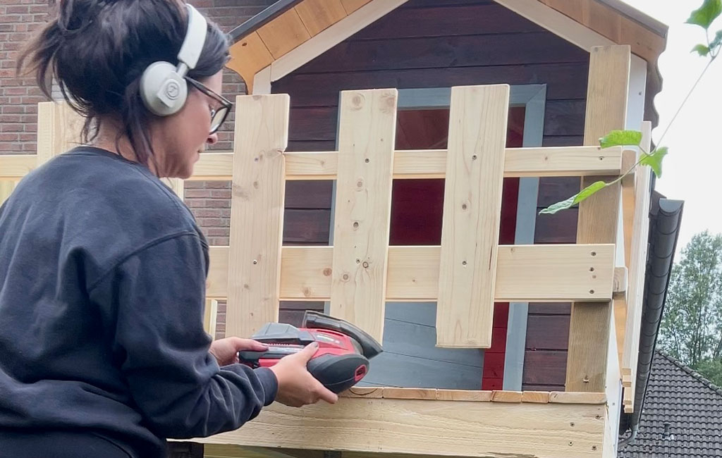 A woman sands the playhouse’s wooden railing with a hand sander.