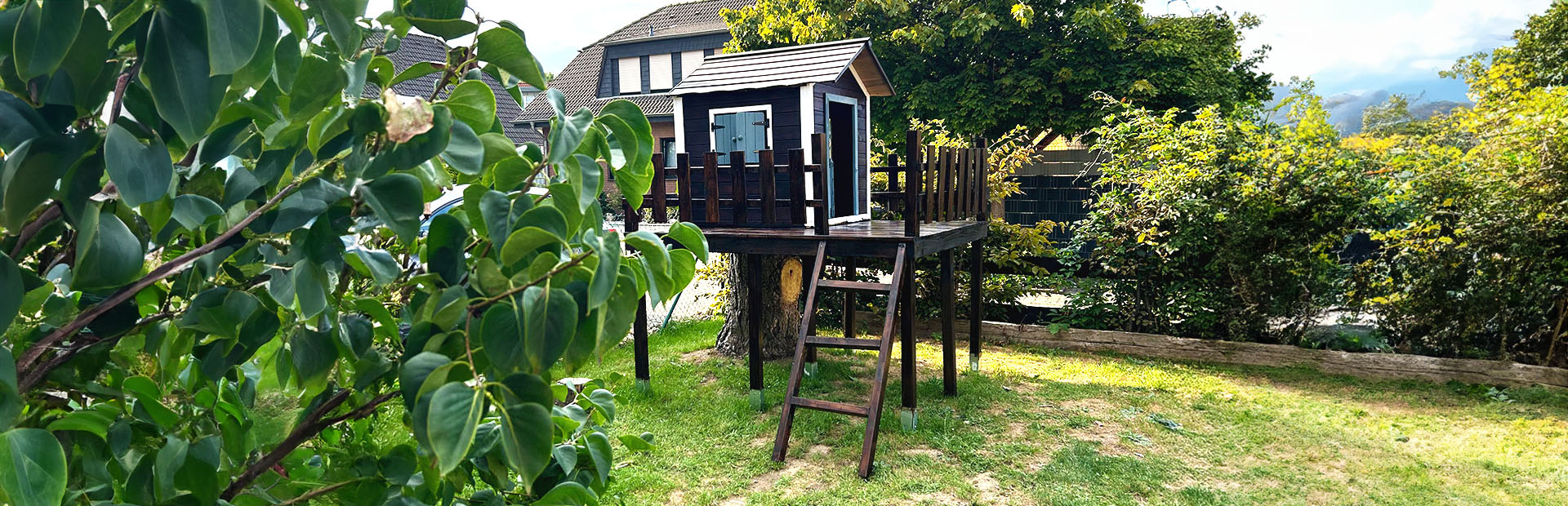 The completed playhouse with wooden railing stands elevated on stilts in the garden next to a tree stump.
