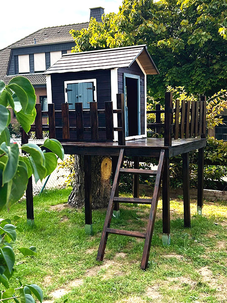 The completed playhouse with wooden railing stands elevated on stilts in the garden next to a tree stump.