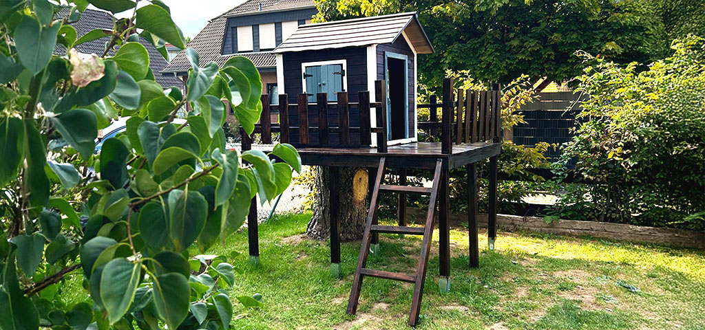 The completed playhouse with wooden railing stands elevated on stilts in the garden next to a tree stump.