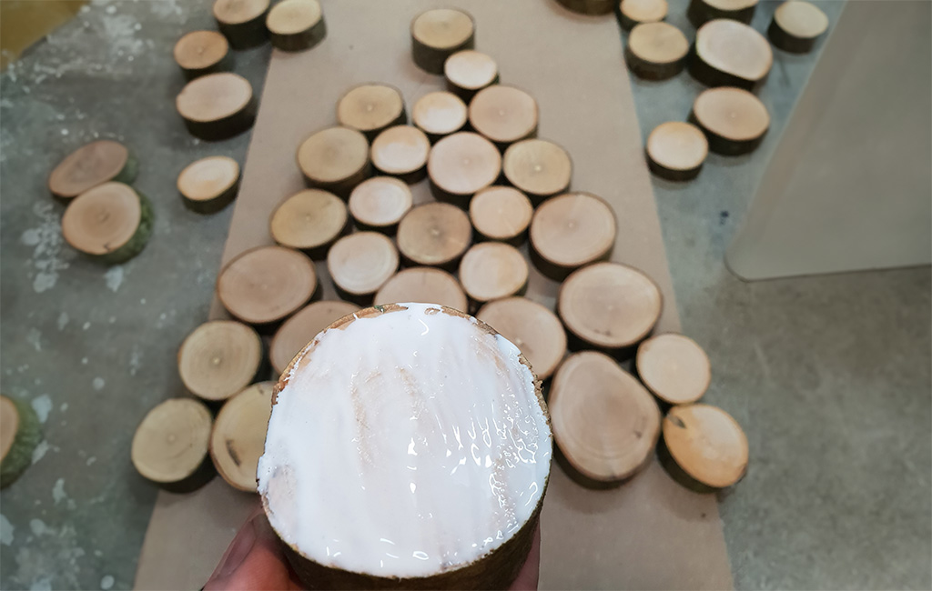A round wooden disc with glue is held in hand, many more discs are laid out on the floor in a tree shape.