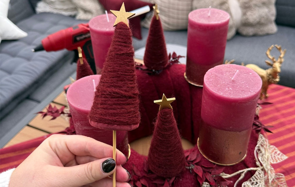 A hand holds a small Christmas tree made of red yarn with a golden star, Advent wreath in the background.