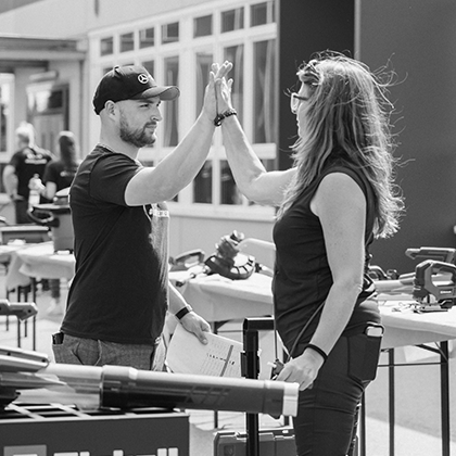 Egy férfi és egy nő high five-ot ad egymásnak egy szabadtéri Einhell-rendezvényen szerszámkiállítással, fekete-fehér fotón.