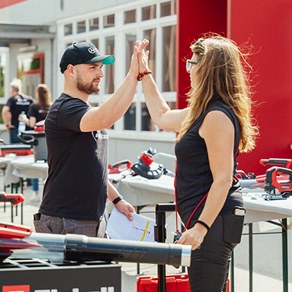 Egy férfi és egy nő high five-ot adnak egymásnak egy szabadtéri Einhell-rendezvényen, ahol szerszámkiállítások vannak.