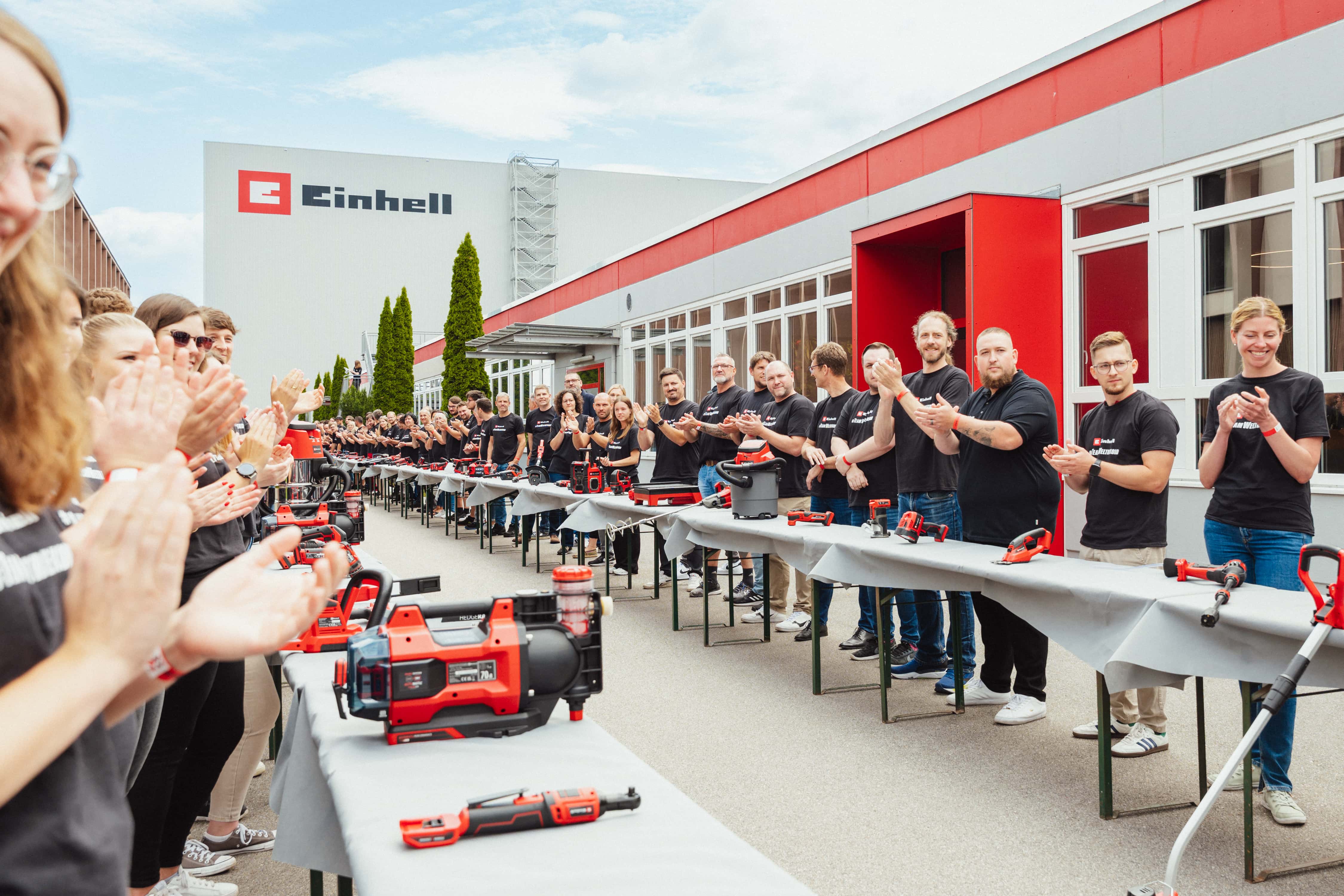 Numerous Einhell employees clap along a long row of tables with cordless tools on the company premises.